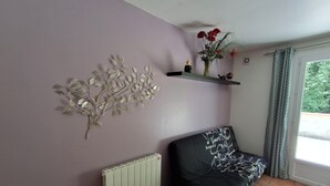 Una sala de estar con un sofá, una mesita redonda pequeña, un radiador, un adorno en la pared, una estantería con flores y una ventana con cortinas.