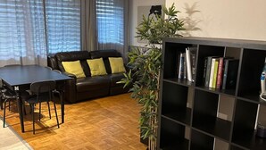 Una sala de estar con un sofá, una mesa de comedor con sillas, una estantería y una planta grande en maceta.