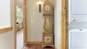 A hallway with a wooden floor, a wall-mounted clock, a light fixture, and a white appliance, likely a washer and dryer. - Maison de Neige Breathtaking Mountain Stay for 8 w/ Hot Tub (Vail)