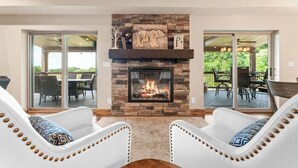 Una chimenea con revestimiento de piedra, dos sillones blancos tapizados con cojines estampados y una zona de patio con mobiliario exterior.