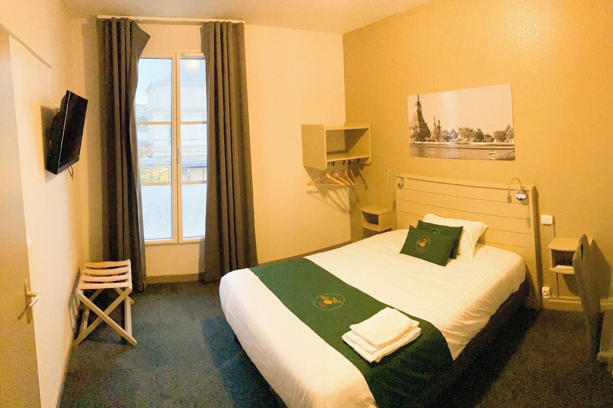 A hotel room with a bed, a chair, a window with curtains, a wall-mounted TV, and a picture of a building.
