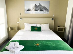 A hotel room with a bed, a green bedspread, white pillows, a green pillow, a wall-mounted picture of pyramids, and a small shelf on the nightstand. - Colette Hotel (Niort)