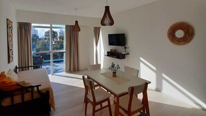 A modern living room with a sofa, dining table, and chairs. There is a TV mounted on the wall and a window with curtains.