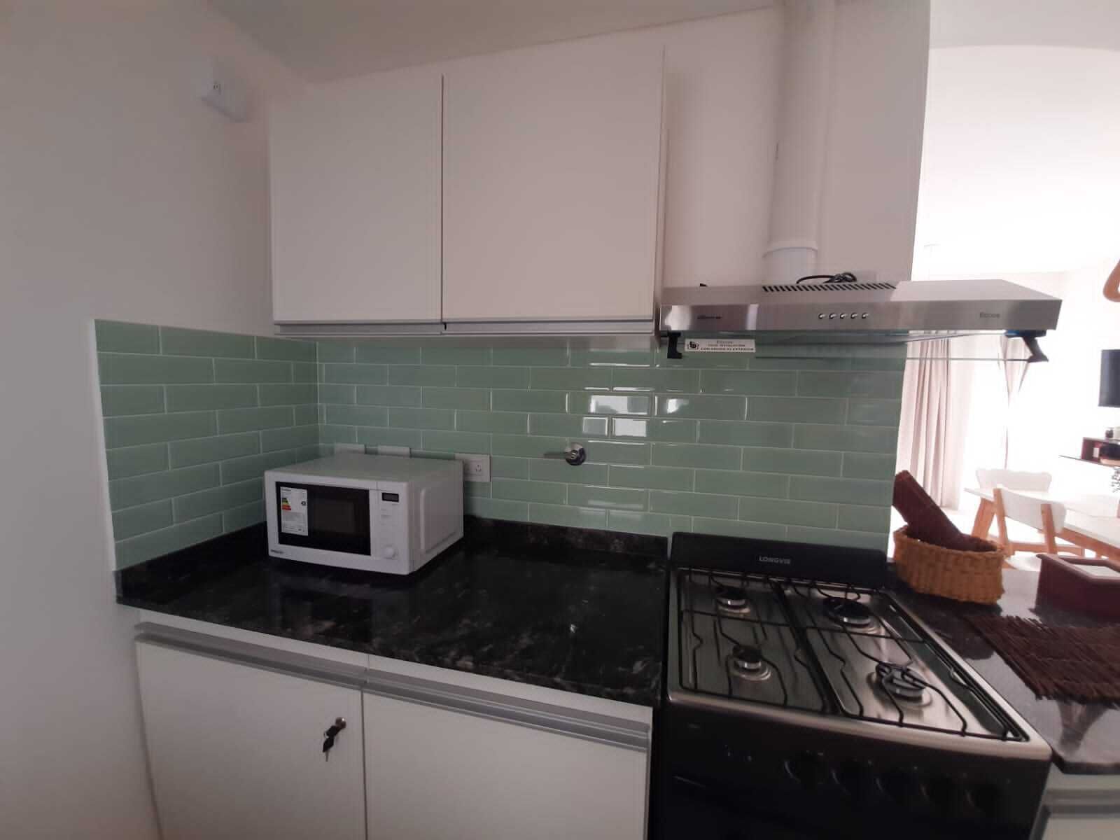 Una cocina compacta con armarios blancos, estufa a gas, microondas y salpicadero de azulejos verdes.