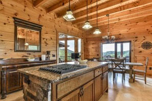 A wooden cabin interior with a kitchen island, granite countertop, wooden cabinets, pendant lights, and a dining area with a table and chairs. - Timber Trail Lodge | Blue Ridge, GA (Blue Ridge)