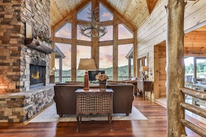 A log cabin interior with a stone fireplace, wooden furniture, and large windows. - Timber Trail Lodge | Blue Ridge, GA (Blue Ridge)