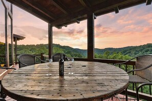 A round wooden table with four chairs, a bottle, and two wine glasses on a veranda with a view of a forested landscape.