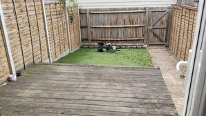 Een achtertuin met een houten terras, een stukje gazon en een metalen poort.