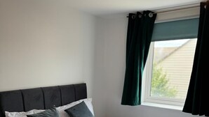 Een net opgemaakt bed met witte lakens en donkere kussens, een donker hoofdbord, en een raam met groene gordijnen.