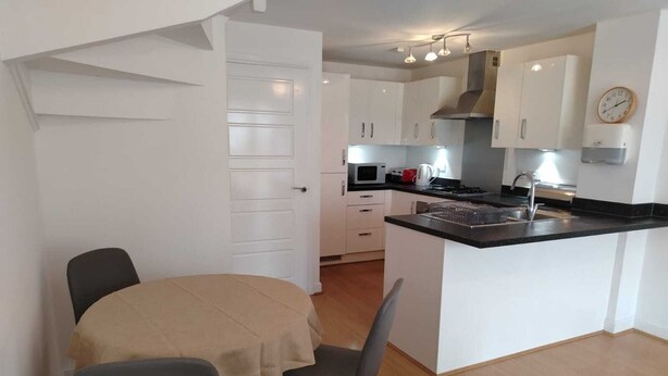 Een compacte keuken met witte kastjes, een magnetron, een fornuis en een spoelbak. Er is een eethoek met een tafel en stoelen.