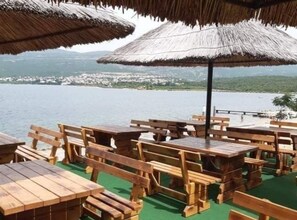 Un restaurant situé en bord de mer, avec des tables et des bancs en bois, des parasols en chaume et une vue sur la mer et les bâtiments éloignés.