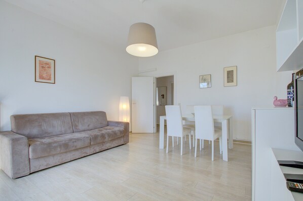 A modern living room with a beige sofa, a dining table with chairs, and a TV on a stand. - Sunny balcony (Venezia)