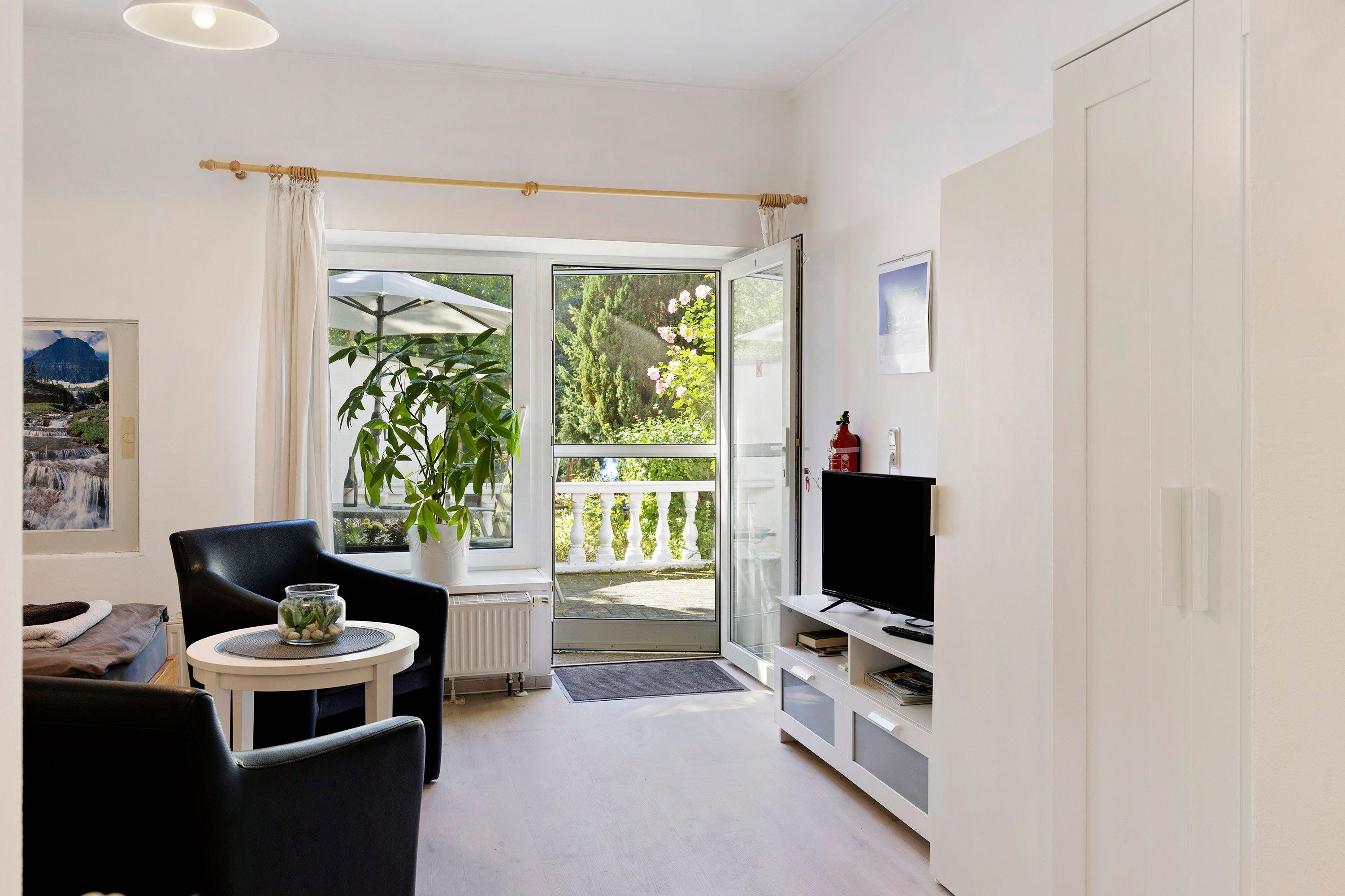 A modern living room with a black chair, a small table with a vase, a TV stand, and a sliding glass door leading to an outdoor area with a plant and a view of a mountain.