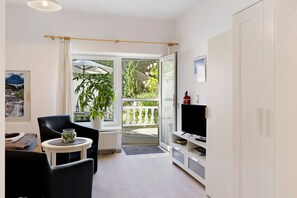A modern living room with a black chair, a small table with a vase, a TV stand, and a sliding glass door leading to an outdoor area with a plant and a view of a mountain. - Apartment 'Behling 1 with Terrace' with Shared Terrace, Communal Garden and Wi-Fi (Liepe)