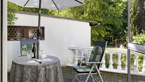 En patio med et bord, stole, en parasol og udsigt over en have.