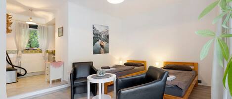 A room with a bed, two chairs, a small table, a radiator, a window with curtains, and a painting on the wall.