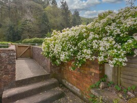 Una escalera de piedra que sube hacia un portón de madera, rodeada de abundante vegetación y flores blancas.