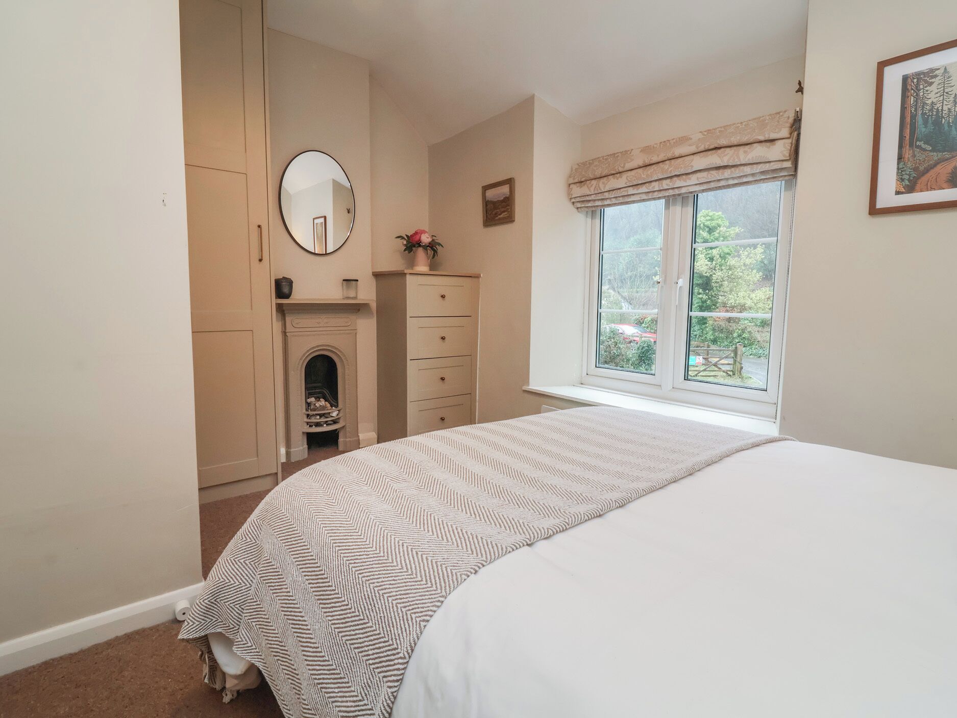A bedroom with a bed, a dresser, a mirror, a fireplace, and a window with a view of greenery.