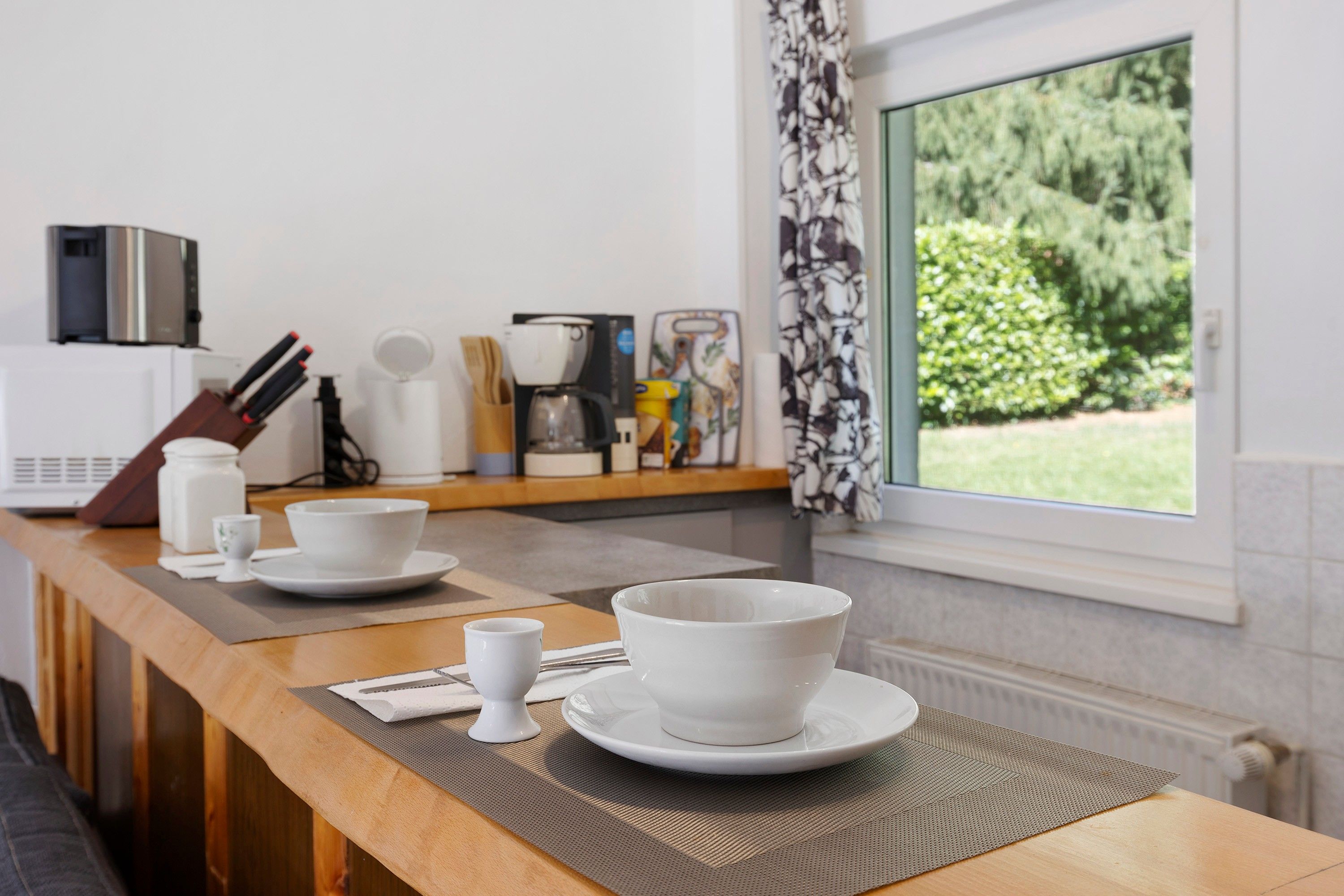 Eine Küche mit einer Holzauslage, weißen Tellern, einer Kaffeemaschine und einem Fenster mit Blick auf Grünflächen.