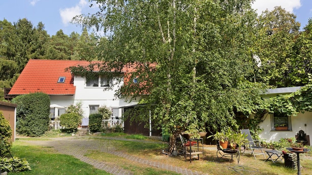 Une maison avec un toit rouge, un chemin pavé et un jardin où poussent différentes plantes, ainsi qu’une brouette.