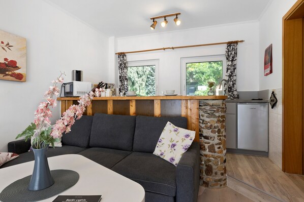 A modern living room with a grey sofa, a white coffee table, and a vase of flowers. There is a kitchen area with a stone wall and a refrigerator.