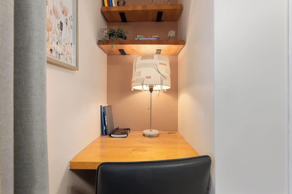 Un bureau d’angle avec une lampe, une chaise, une étagère avec des livres et des objets décoratifs, et un tableau encadré accroché au mur.