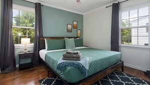 Une chambre à coucher comprenant un lit, une table de chevet, une lampe, un ventilateur de plafond et une fenêtre avec des rideaux.