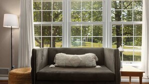 Un canapé gris foncé avec un coussin blanc, placé devant une grande fenêtre à carreaux, un lampadaire et une petite table en bois.