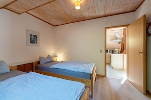 A room with two beds, a wooden ceiling, a framed picture on the wall, and a kitchen area visible through an open door.