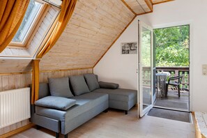 Una habitación con techo inclinado, un sofá gris, suelo de madera y una puerta que da a un área exterior con mesa y sillas.