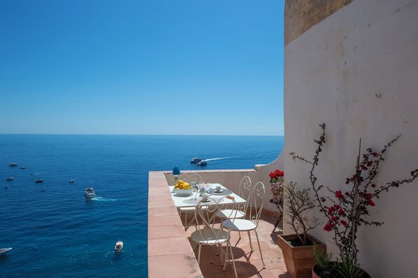 A terrace with a table set for dining, overlooking a sea with boats, and potted plants on the side.