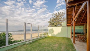 Ein modernes Haus mit Glaswand, das auf einen Strand blickt, einer Holzterrasse und einem grünen Rasen.