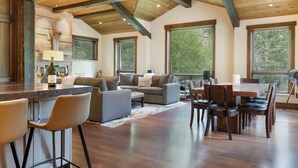 A spacious living area with a wooden ceiling, a bar area with stools, a dining table with chairs, and large windows offering a view of the outdoors. - Basingdale Hollow - For 10 (Vail)