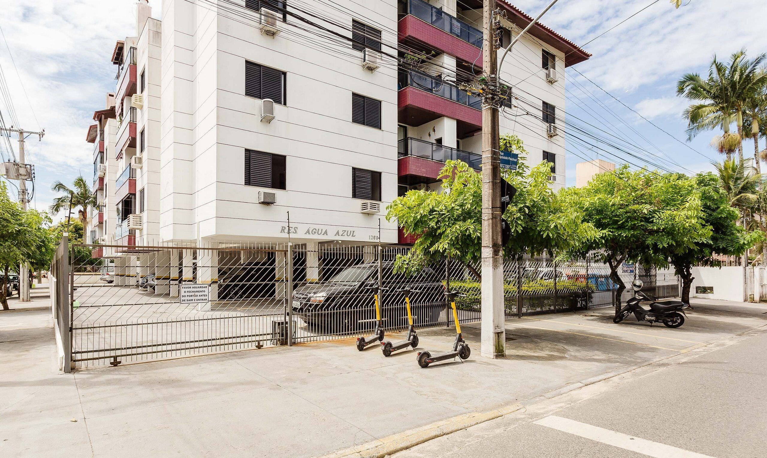 A multi-story residential building with balconies, a gated parking area, and a scooter parked outside.