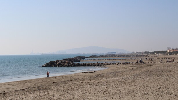 Una playa con formaciones rocosas, una persona de pie junto al agua y edificios al fondo.