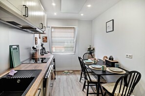 Une cuisine compacte dotée d’appareils électroménagers modernes, un coin repas avec une table pour deux personnes et une fenêtre avec des stores.