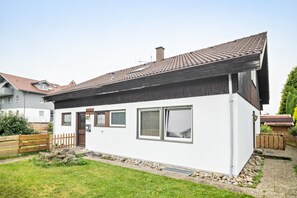 Une maison moderne à deux étages, avec une façade blanche, un toit foncé et un petit jardin à l’avant, agrémenté d’une clôture en bois et d’un chemin en pierre.