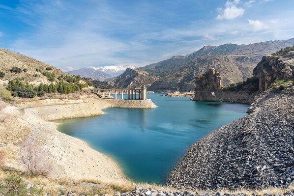 Una presa con un puente, agua tranquila, terreno rocoso y montañas al fondo.