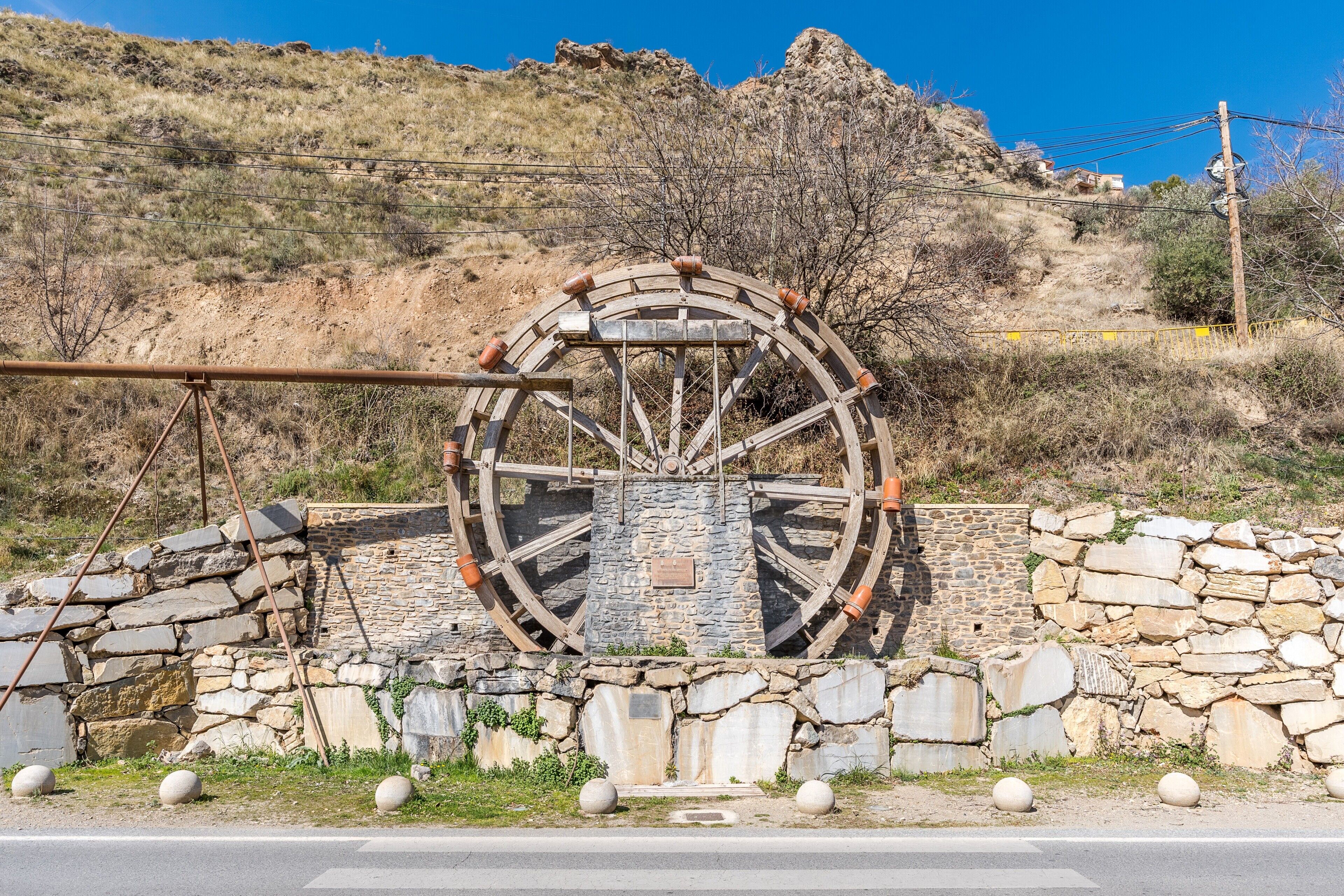 Una gran estructura de noria con muros de piedra y estructura metálica, ubicada frente a un paisaje montañoso.