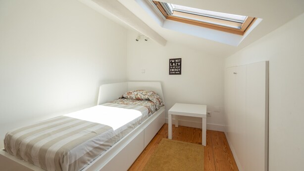 A small, well-lit bedroom with a skylight, a bed with striped bedding, a small white bedside table, and a wall-mounted light fixture. - Bairro Alto Tiny Studio by Homing (Lisbon)