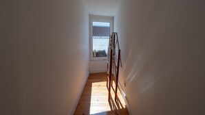 A narrow hallway with wooden flooring and white walls, leading to a window with a view of a building. - Bairro Alto Tiny Studio by Homing (Lisbon)