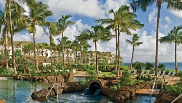 Een zwembad van een resort met helder water, palmbomen en uitzicht op de oceaan.