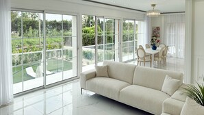 Un salón amplio con un gran ventanal corredera que da a un jardín, un sofá blanco y una zona de comedor con sillas y una mesa.