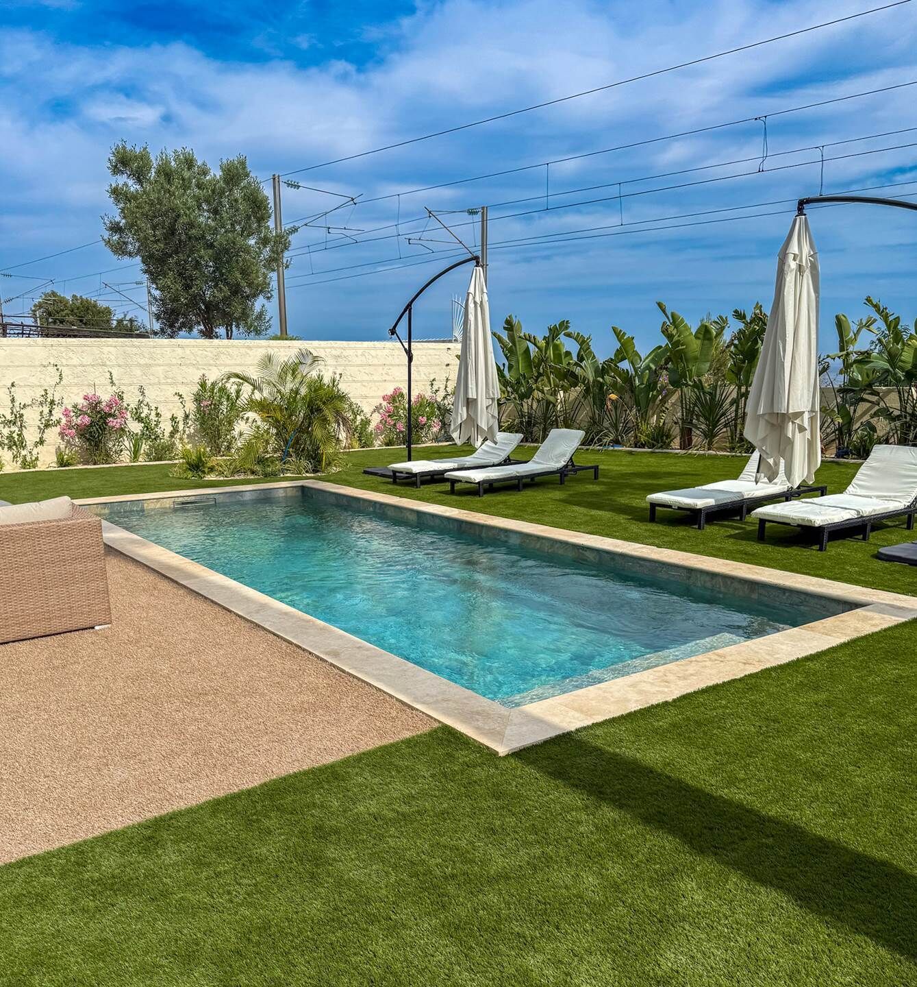 A rectangular swimming pool with two lounge chairs and umbrellas, surrounded by artificial grass and tropical plants.