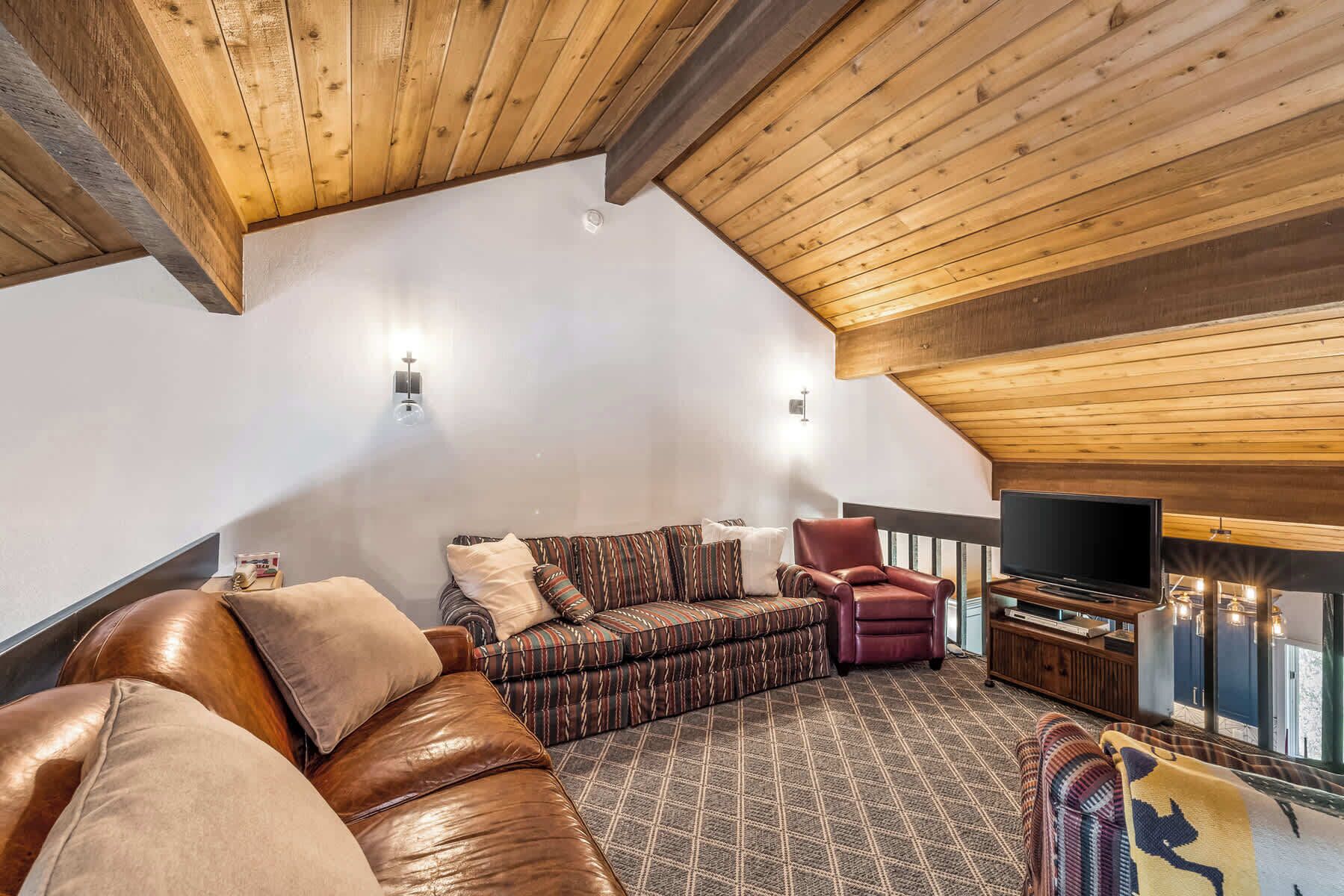 A cozy loft-style living room with a high ceiling, a leather sofa, a patterned armchair, a red armchair, a television, and a fireplace.