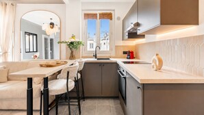 A compact kitchen with a dining area, a window with blinds, and a view of buildings outside. - 1536 Daumesnil (Paris)