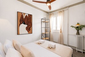 Habitación de hotel con cama, estante en la pared, ventilador de techo, ventana con cortinas y radiador.
