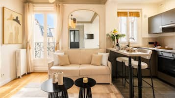 A modern apartment with a living room, kitchen, and dining area. There is a sofa, two round tables, and a kitchen counter with stools.