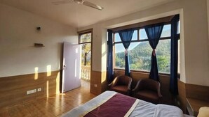 A hotel room with a bed, two chairs, a ceiling fan, a window with curtains, and a door leading to a balcony. - Villa Kartina 3 BHK near Sattal Lake (Bhowāli)
