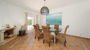 Un coin repas avec des chaises en osier, une table en verre, un luminaire suspendu et un tableau représentant une vague accroché au mur.